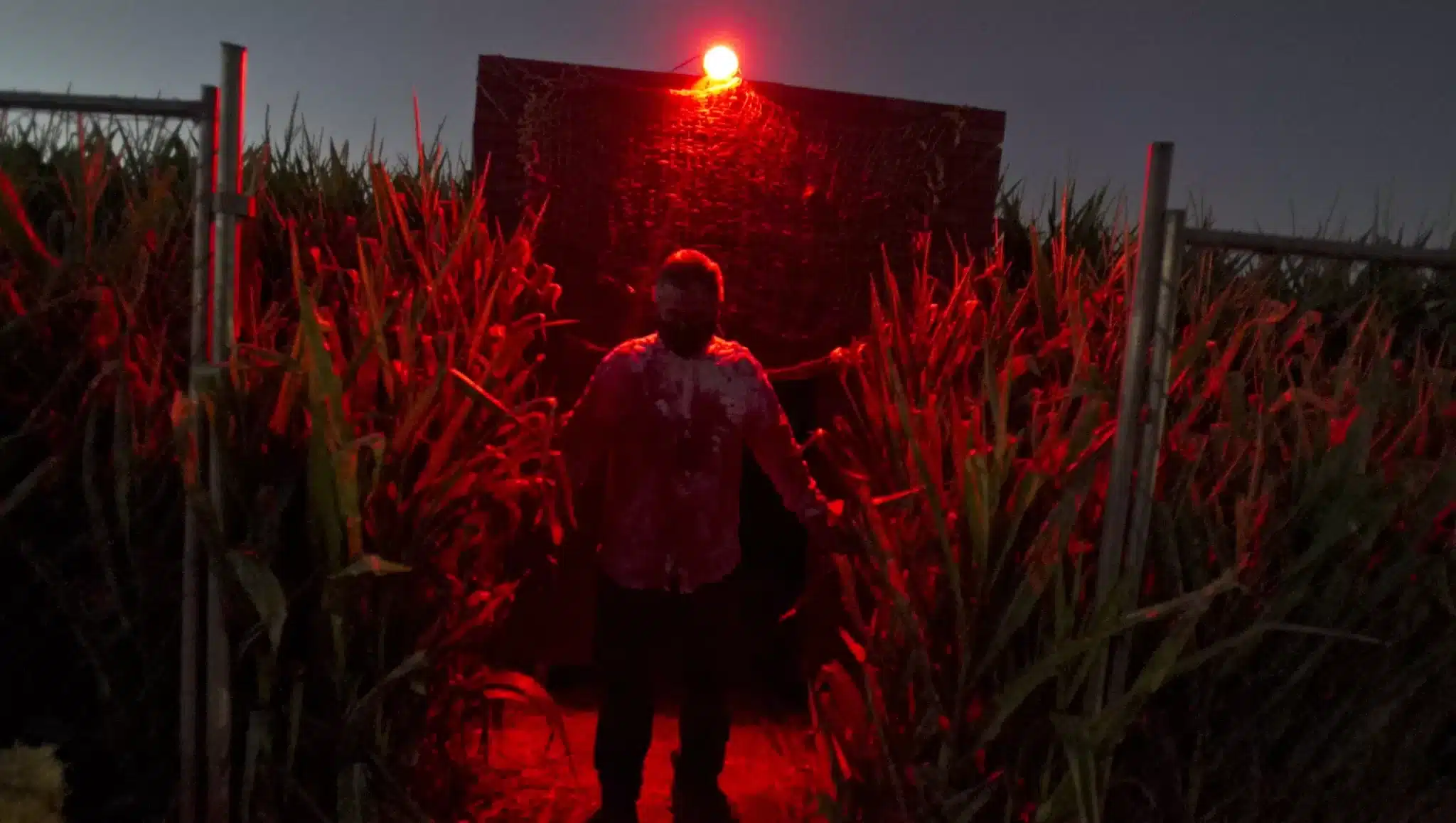 Character Covered in Blood Standing in Field with Red Light Behind Him at Terror in the Corn