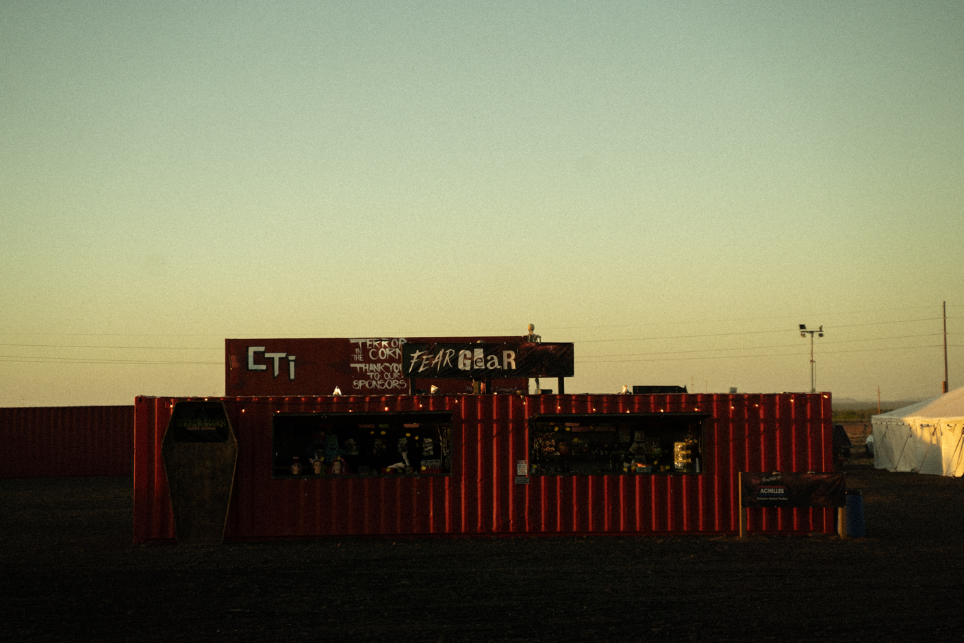 Red corrugated-storefront container at sunset with two display windows selling merchandise and a 'FEAR Gear' sign above it in a desert setting
