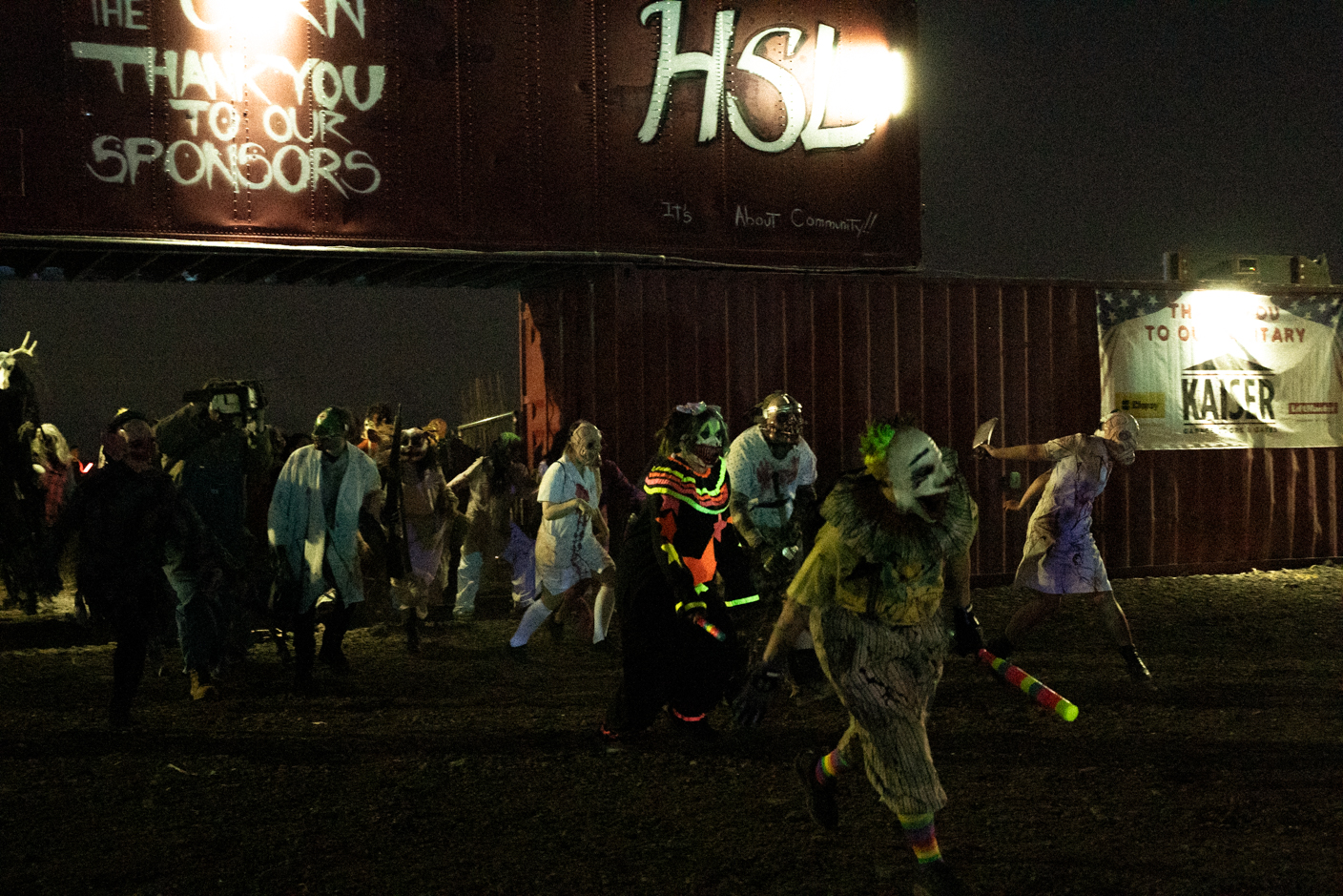 Nighttime zombie walk with people in tattered, colorful costumes marching past a large banner and metal shipping container in the background.