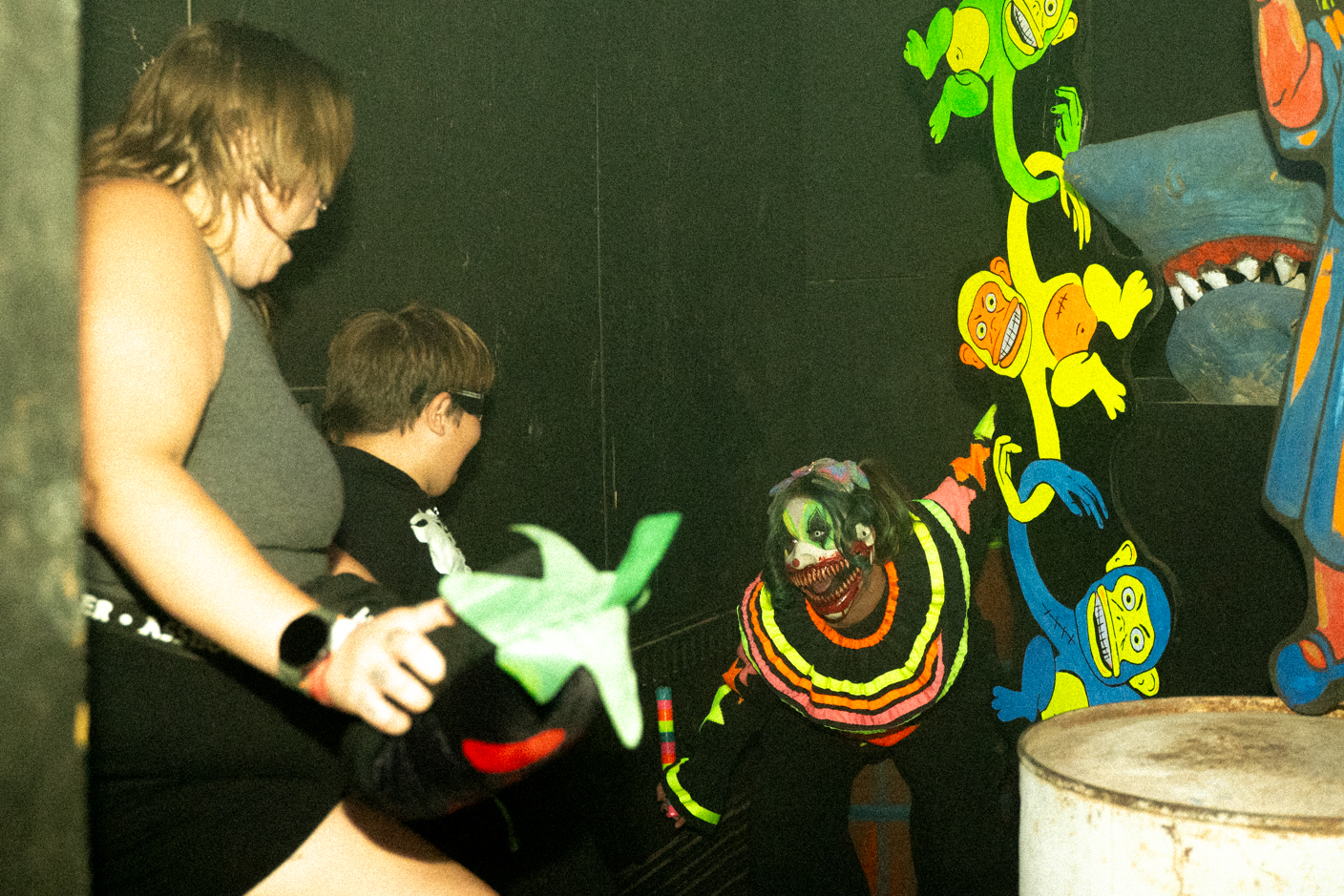 Person in a neon clown costume with green and orange makeup moves toward the camera in a dark, mural-decorated room.