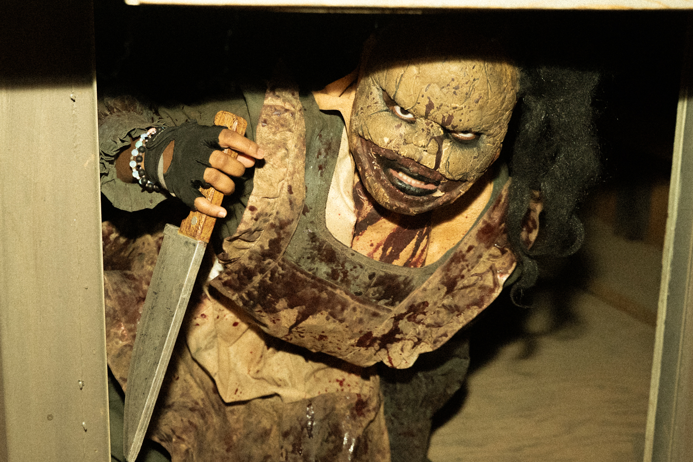 A scary masked figure with a cracked mask and blood-stained clothes, holding a large knife and peeking through a doorway.