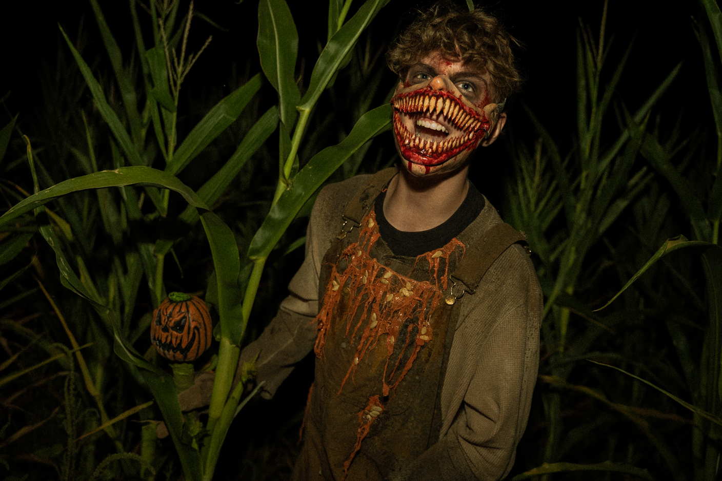 Person in a tattered brown costume and a large red, toothy mask; stands in a dark cornfield with a small carved pumpkin prop.