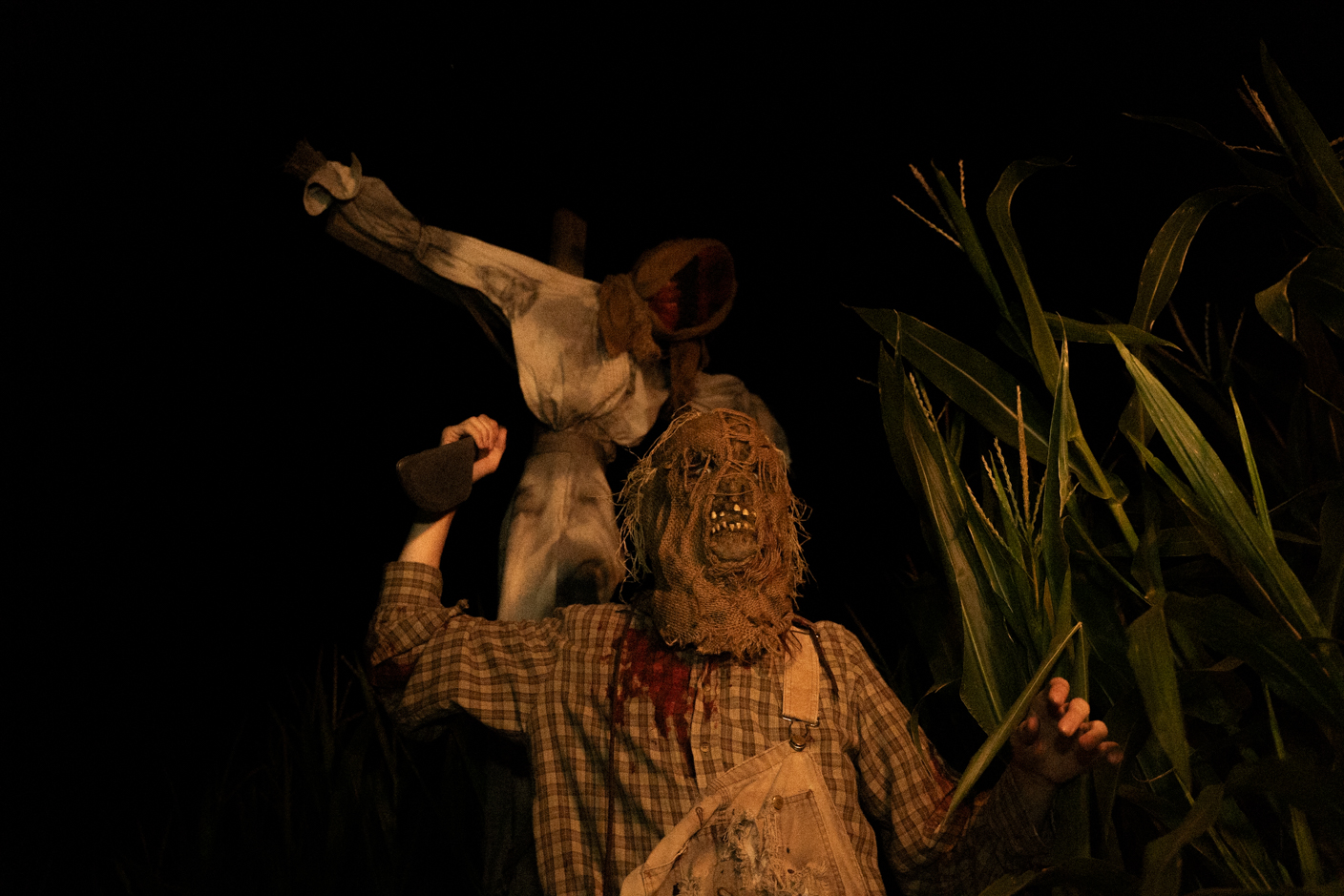Dark outdoor scene of two creepy scarecrow figures in a cornfield at night; one wears a burlap mask and tattered clothing, the other raises an axe above its head.