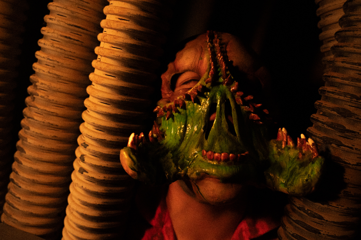 Person wearing a green dragon mask with red spikes peeking between stacked beige coiled columns in dim lighting
