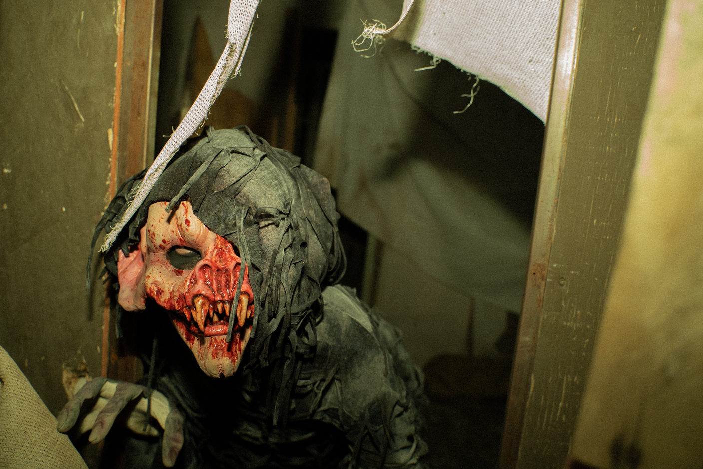 Ragged horror mask with a red, blood-streaked face and long fangs peeking from a doorway.