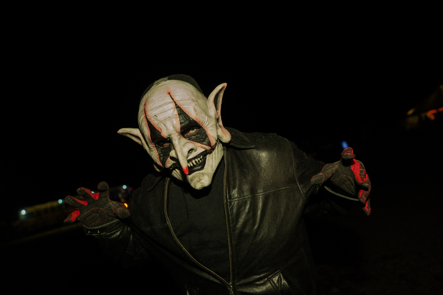 Person in a pale, horror-masked costume with pointed ears and red-black makeup, wearing a black leather jacket at night, reaching toward the camera.